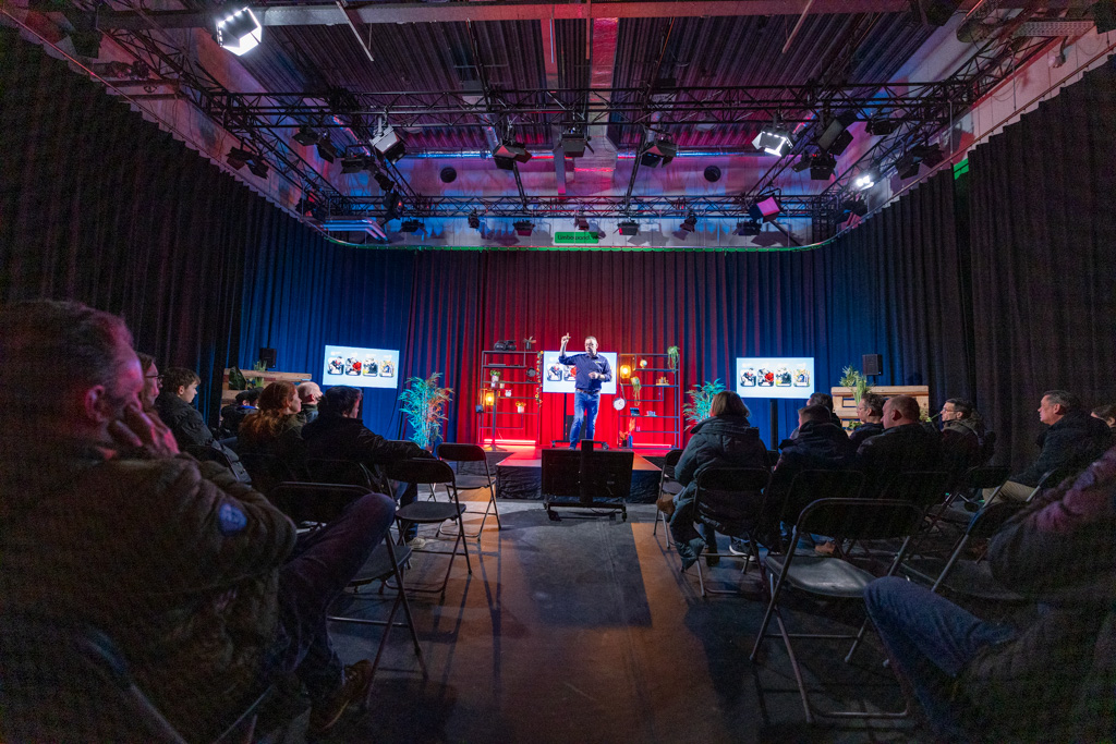Presentator op podium die kijkt naar publiek met kleurrijke lampen op de achtergrond. 