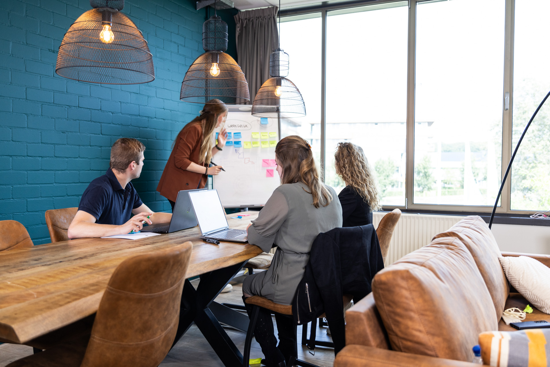 Een groep collega's werkt samen tijdens een brainstormsessie in een lichte vergaderruimte. Een vrouw in een bruin jasje schrijft op een flip-over met het thema 