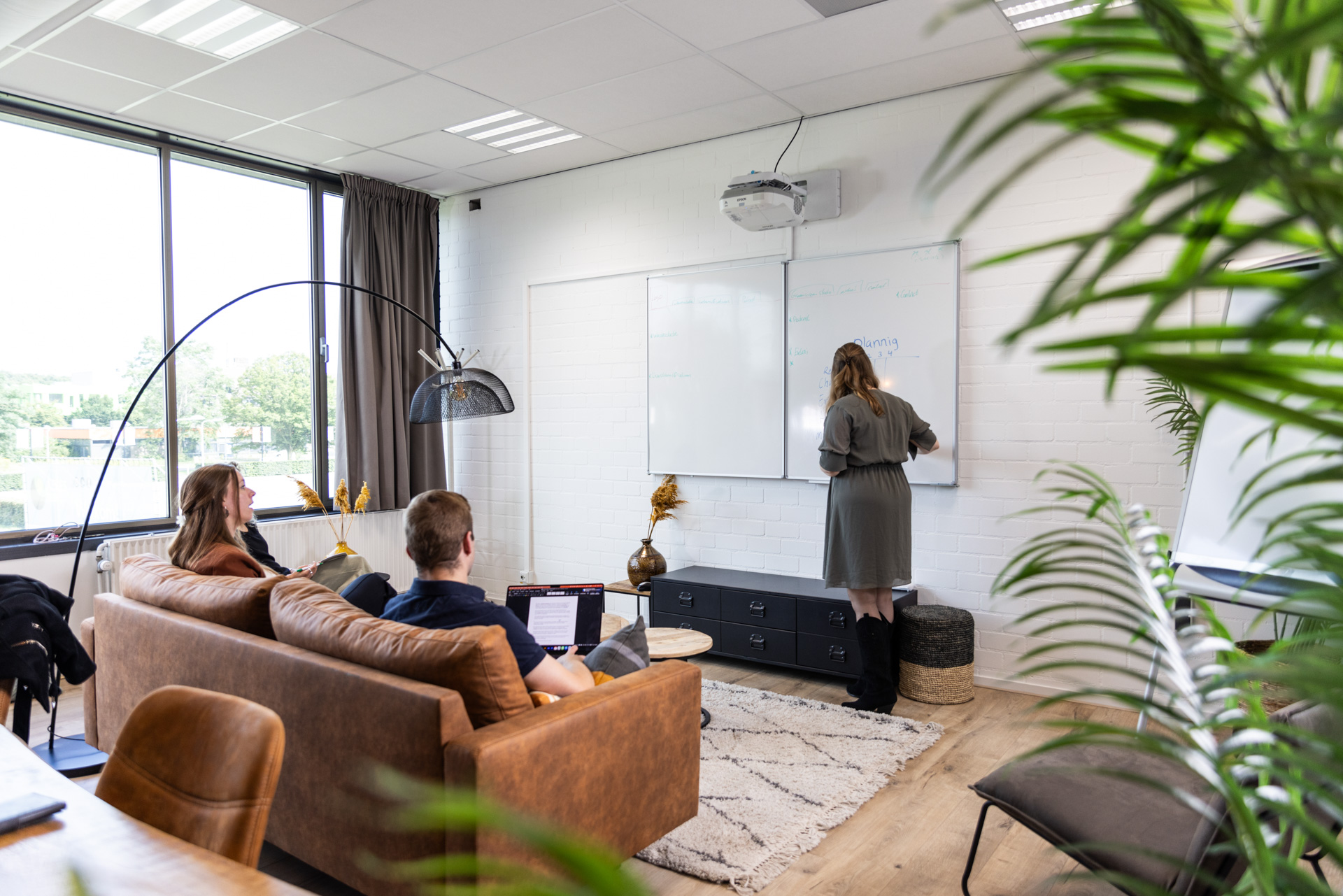 Een vrouw schrijft op een whiteboard terwijl twee collega's ontspannen op een bruine leren bank meedenken. De ruimte is modern en sfeervol ingericht met grote ramen, een houten vloer en groen decor. Een laptop staat open met notities, en een projector hangt aan het plafond. De ontspannen setting bevordert creativiteit en samenwerking.