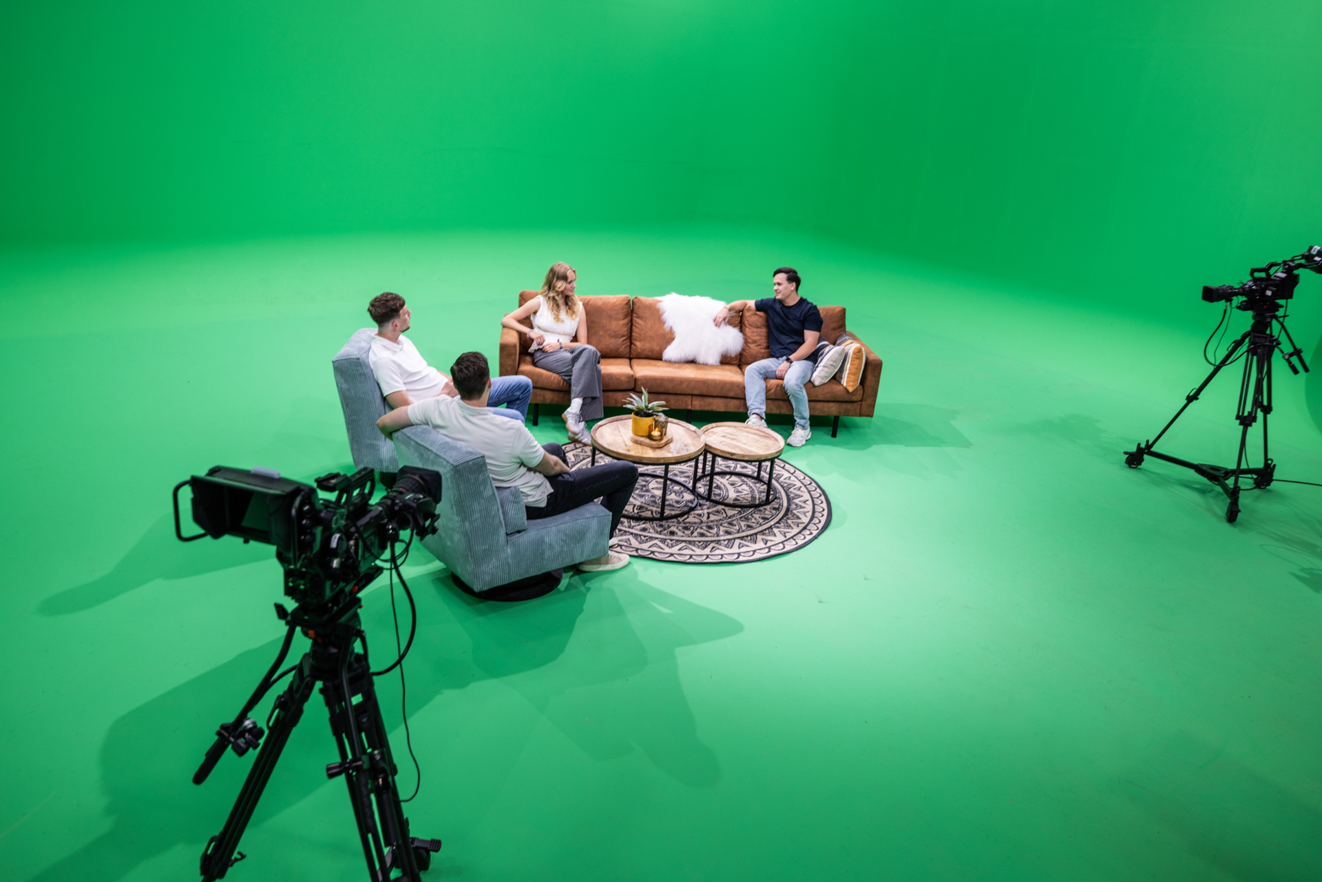 Vier personen zitten in een informele talkshow-opstelling in een ruime green screen studio. Ze zitten op een bruine bank en twee grijze fauteuils rond een set ronde tafels met decoratieve elementen. De camera-opstelling met meerdere professionele videocamera’s op statieven is zichtbaar op de voorgrond, waarmee de opnames professioneel worden vastgelegd. De groene achtergrond maakt het mogelijk om digitale achtergronden toe te voegen tijdens de nabewerking.