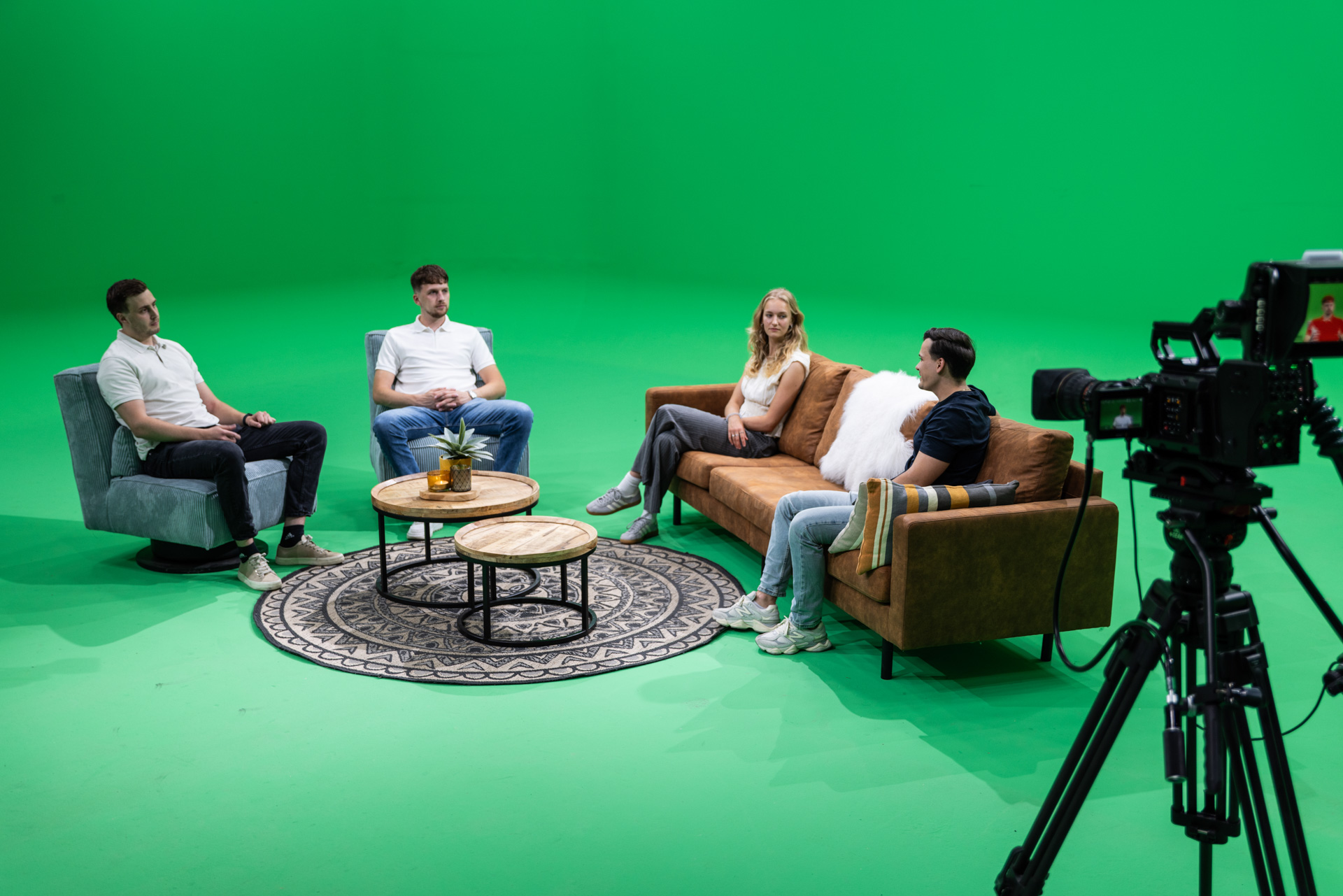 Vier personen zitten in een informele gespreksopstelling op een bruine bank en grijze fauteuils, terwijl een grote videocamera op statief de scène vastlegt. De opstelling omvat drie ronde houten tafels met een plant en decoratieve kaarsen op een stijlvol zwart-wit kleed. De groene achtergrond biedt mogelijkheden voor digitale nabewerking en het invoegen van virtuele decors.